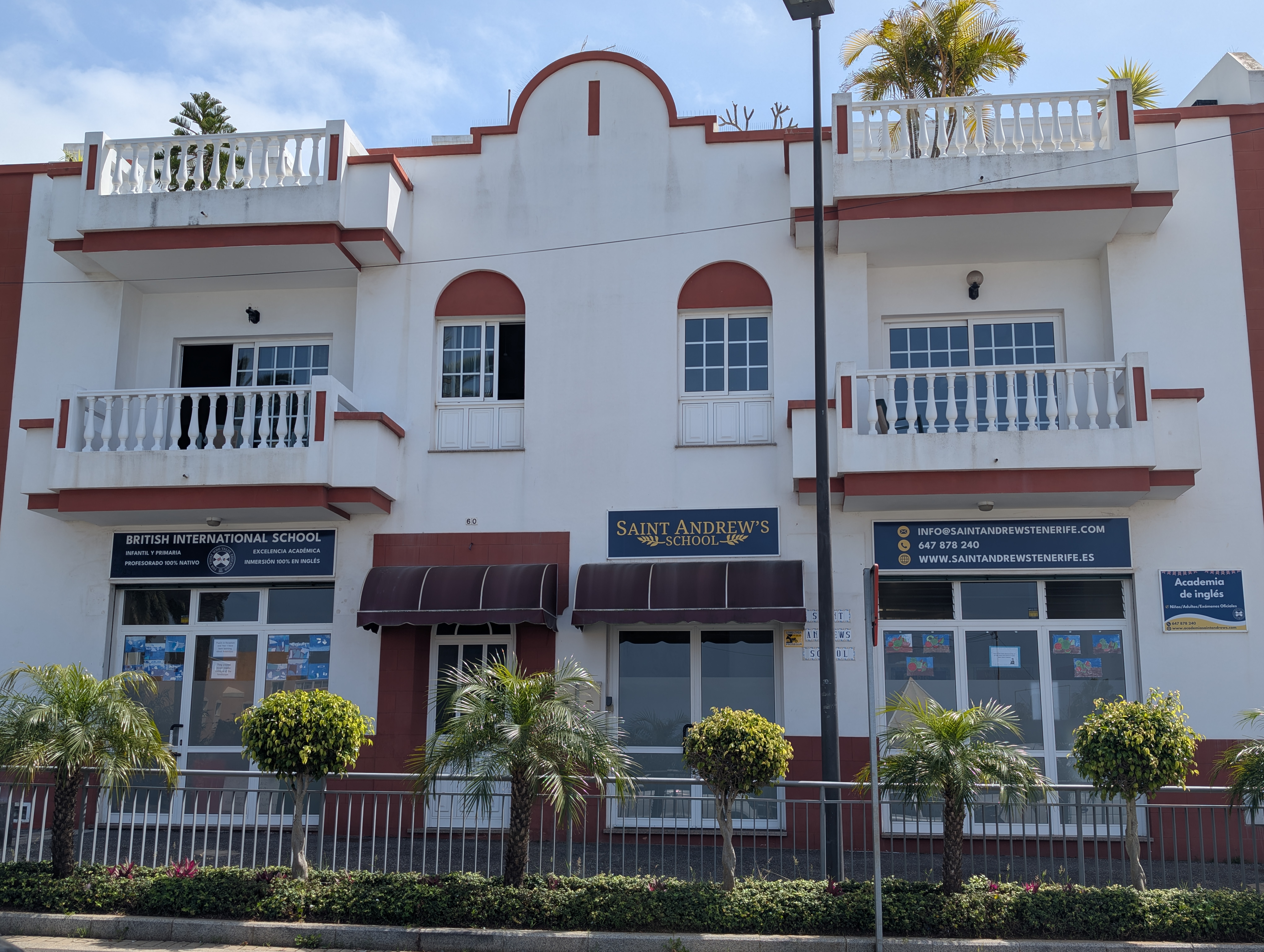 Fachada del Saint Andrew’s School, un colegio británico internacional con detalles arquitectónicos blancos y granates, palmeras decorativas en la entrada y carteles informativos sobre sus servicios educativos.