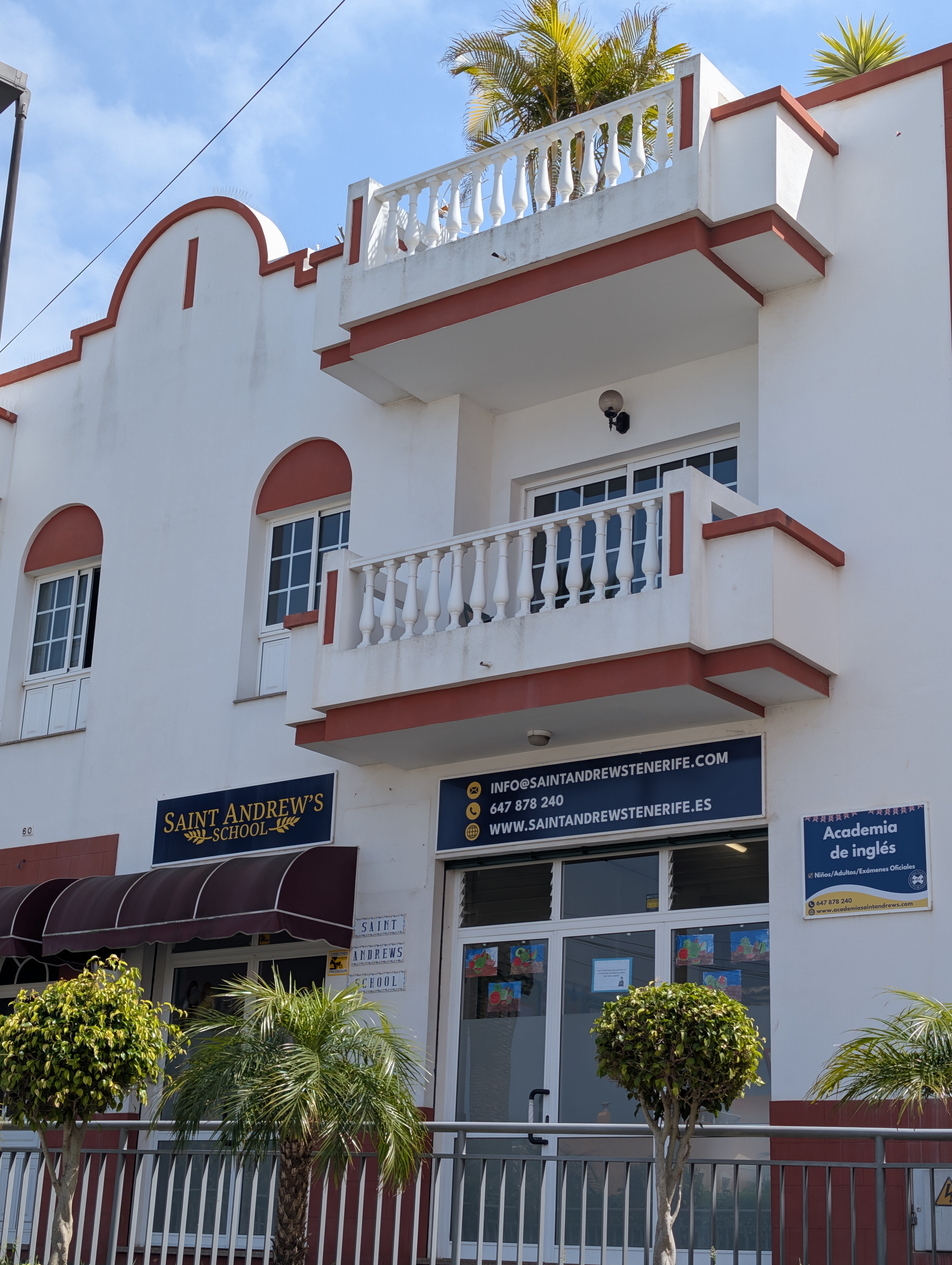 Fachada del Saint Andrew’s School, un colegio británico internacional con detalles arquitectónicos blancos y granates, palmeras decorativas en la entrada y carteles informativos sobre sus servicios educativos.